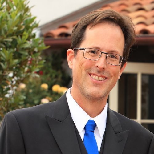 Headshot of Scott Seward. Wearing a dark suit, white shirt, bright blue tie. He wears glasses and a small. He's outside a building with a tile roof.